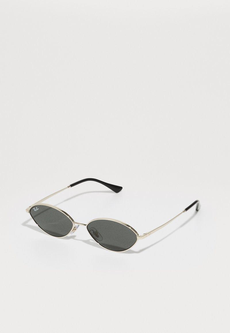Gafas de sol ovaladas con lentes grises, montura de metal dorado delgada, puntas de patilla negras y un diseño minimalista. Presenta el logotipo en la lente.