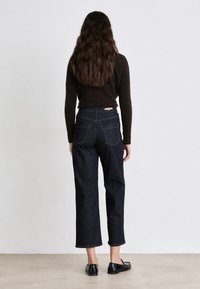 Dark denim jeans with a straight leg design and raw hem, paired with a dark knit sweater and black slip-on shoes, viewed from behind.