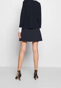 Femme debout contre un mur, portant une jupe marine, un pull assorti avec des poignets blancs, et des chaussures à talons noires avec bride à la cheville, vue de dos.