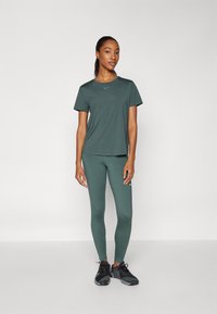 Mørkegrønn treningstrøye og matchende tights, laget av stretch-stoff, med en subtil logo-detalj, sammen med svarte joggesko.