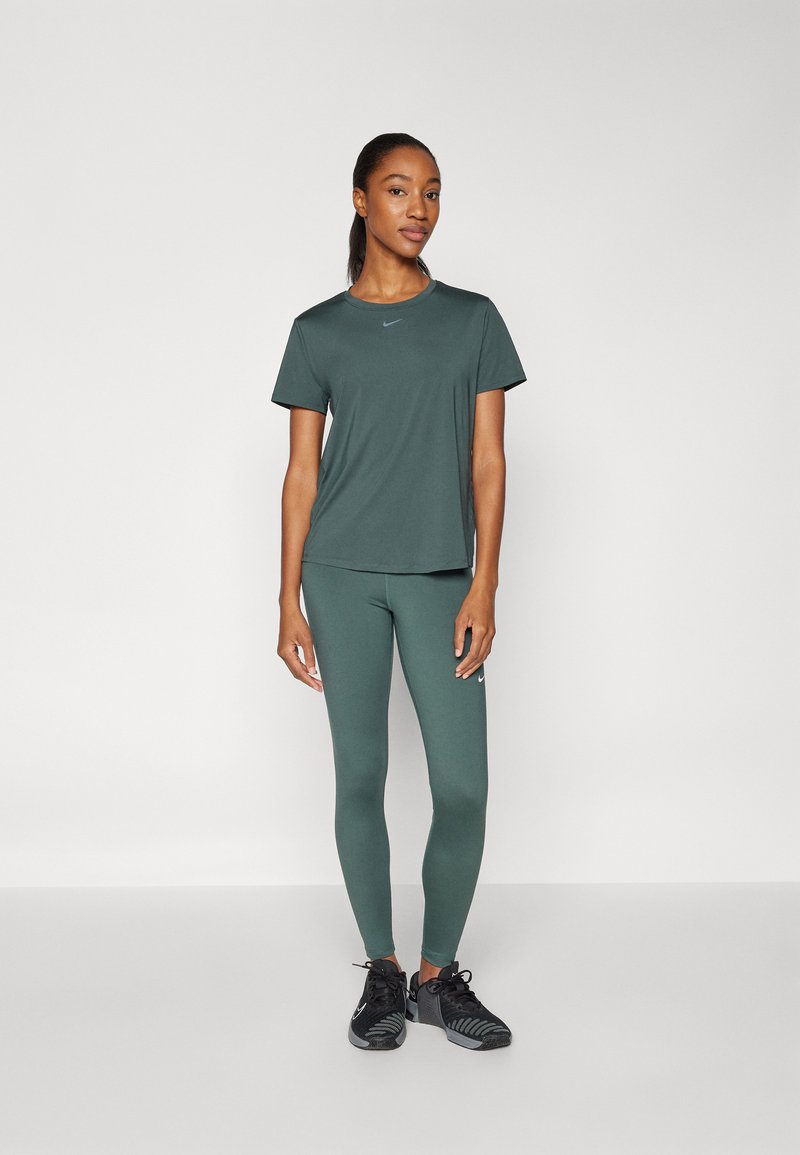 T-shirt de sport vert foncé et leggings assortis, fabriqués en tissu extensible, avec un logo discret, associés à des chaussures de sport noires.
