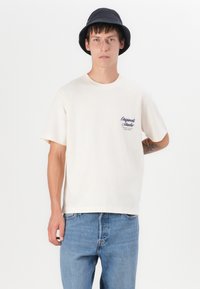 T-shirt crème à manches courtes avec un texte graphique bleu sur la poitrine gauche, associé à un jean en denim bleu clair et un chapeau seau noir.