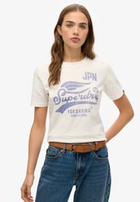 Hvid bomulds t-shirt med blå grafisk tekst "Superdry", med designs inklusiv "JPN", et vinge motiv og "Yokohama". Båret med blå jeans.