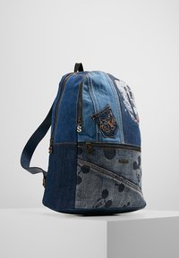 Džínový batoh v různých odstínech modré, který má přední kapsu s motivem Mickey Mouse a dvě přihrádky na zip. Zdobený šicími detaily.