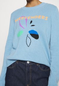 Pull en tricot bleu clair avec des formes abstraites multicolores et le texte "WEEKENDERS" en orange. Poignets et ourlet côtelés.