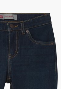 Jeans skinny de denim azul oscuro con cierre de cremallera, botón plateado y dos bolsillos delanteros. Costuras amarillas a lo largo de las líneas y bordes de los bolsillos.