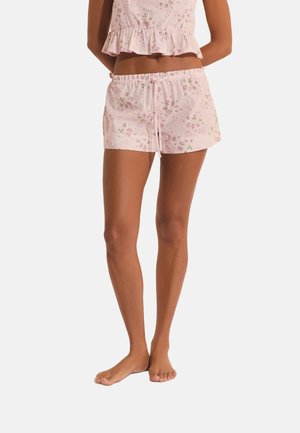 Femme portant un short de pyjama rose clair à motifs floraux avec cordon de serrage et un haut assorti à ourlet à volants, debout pieds nus sur fond blanc.