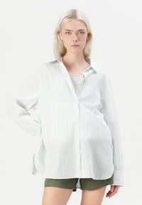 Camicia bianca a maniche lunghe con righe azzurre, con bottoni sul davanti e vestibilità rilassata, indossata sopra un top grigio testurizzato e pantaloni corti verdi.