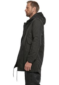 Parka negra con capucha, presenta un cierre frontal con cremallera y broches de presión, cordones ajustables y bolsillos laterales. Tejido texturizado.