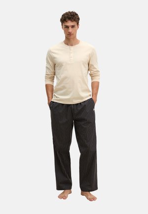 Jonge man staat blootsvoets, draagt een crèmekleurig henley-shirt met lange mouwen en zwarte losse broek met fijne strepen, handen in de zakken.