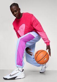 Pink struktureret sweatshirt med et stort design, blå og lilla farveblokerede sweatpants og hvide atletiske sko, som holder en orange basketball.