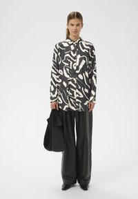 Naine, kes kannab must-valget abstraktse mustriga pluusi, laiu musti pükse, teravnurkseid kingi ja hoiab suurt musta käekotti, seisab valgel taustal.