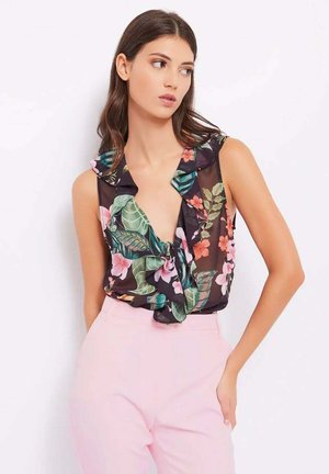 Donna con lunghi capelli scuri che indossa una blusa nera senza maniche a fiori e pantaloni a vita alta rosa chiaro, in posa con una mano in tasca.