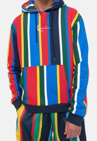 Wielokolorowa bluza z kapturem w paski, obejmująca czerwone, niebieskie, zielone, żółte i białe pasy. Posiada przednią kieszeń oraz kaptur ze sznurkiem.