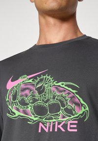 Grå bomulds t-shirt med et grønt og pink grafisk logo, inklusive et stiliseret væsen design og "NIKE" tekst på forsiden.