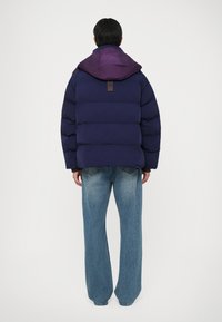Vijolična in temno modra puffer jakna s kapuco, ki ima oznako na hrbtni strani, v kombinaciji s svetlo modrimi širokimi kavbojkami.