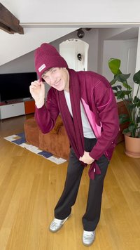 Bordó kötött sapka és hozzáillő kardigán rózsaszín részletekkel, fehér póló, fekete nadrág és ezüst cipő, beltéri növények között pózolva.