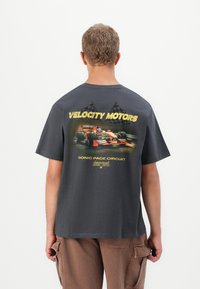 Bărbat îmbrăcat cu un tricou gri închis, având un grafic cu o mașină de curse roșie și steaguri în carouri, cu textul "Circuitul Sonic Pace Velocity Motors" pe spate.