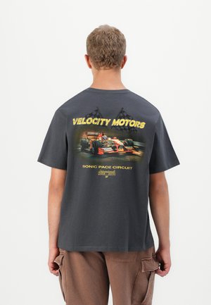 Man med mörkgrå T-shirt med röd sportbil och rutiga flaggor som grafiskt motiv, "Velocity Motors Sonic Pace Circuit" text på baksidan.