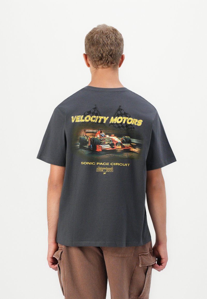 Bărbat îmbrăcat cu un tricou gri închis, având un grafic cu o mașină de curse roșie și steaguri în carouri, cu textul "Circuitul Sonic Pace Velocity Motors" pe spate.