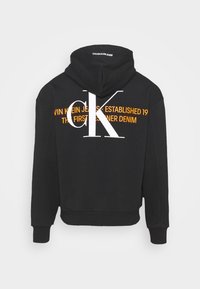 Černá mikina s kapucí, která má na zádech velké bílé logo "CK" a oranžový text. Vyrobena z měkké látky, s kapsou ve stylu klokánka a stahovací kapucí.