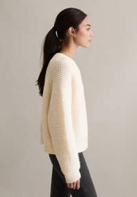 Gruby, kremowy sweter dziany z teksturowanym wzorem w zygzaki, okrągłym dekoltem i ściągaczami na rękawach, zestawiony z czarnymi spodniami, widziany z boku.