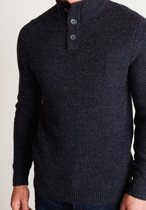Pull gris foncé en tricot avec motif texturé et col côtelé à deux boutons porté par une personne debout, les bras détendus.