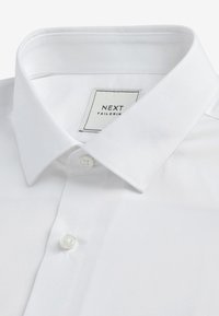 Cămașă albă din bumbac cu guler clasic, închidere cu un singur buton și un etichetă care afișează "NEXT TAILORING". Textură netedă, simplă.