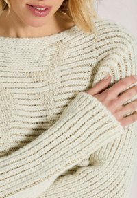Frau trägt einen cremefarbenen grob gestrickten Pullover mit strukturierten horizontalen und diagonalen Rippmustern, die Hand ruht auf der gegenüberliegenden Schulter.