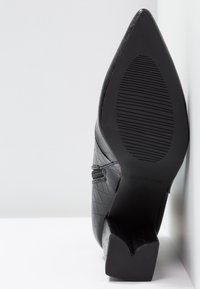 Chaussure noire à talon haut avec un bout pointu, en cuir texturé. Le talon est épais avec un motif antidérapant sur la semelle.