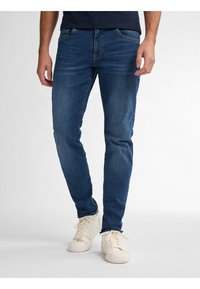 Jeans de mezclilla azul oscuro con corte ajustado, que cuentan con cinco bolsillos y un ligero desvanecimiento. Combinados con zapatillas blancas para un aspecto casual.