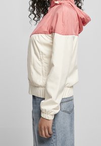 Kurtka w stylu color-blocking z różową górną częścią i kremową dolną częścią, uszyta z gładkiego materiału, z regulowanym kapturem i elastycznymi mankietami.