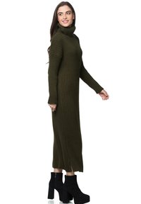 Strickkleid mit Rollkragen in Olivgrün, geripptem Muster, langen Ärmeln und Seitenschlitzen. Kombiniert mit schwarzen Stiefeletten mit Absatz.