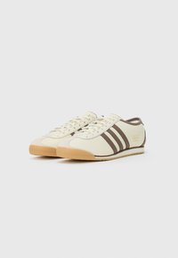 Scarpe da ginnastica beige chiaro con strisce marroni, tomaia in tessuto liscio, suola in gomma e branding sottile sul lato.