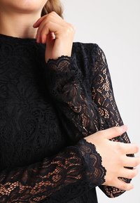 Kvinna iförd en svart spetstopp med lång ärm, händerna som försiktigt rör vid hennes ansikte och motsatta arm mot en enkel grå bakgrund.