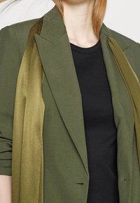 Žena v olivově zeleném blejzru přes černý žebrovaný top, s červenými vlasy částečně viditelnými na jednobarevném pozadí.