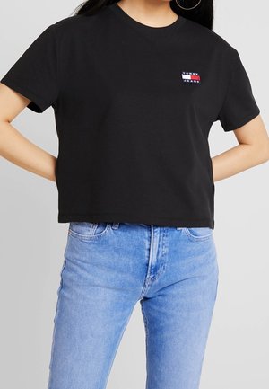 Kobieta w czarnej krótkiej koszulce Tommy Jeans z logo na piersi oraz jasnoniebieskich jeansach z wysokim stanem, ręce na biodrach.