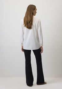 Bela srajca z dolgimi rokavi in napihnjenimi rameni, kombinirana z črnimi razširjenimi hlačami. Gladka tekstura; srajca ima zaobljen rob in zapenjanje na manšetah.