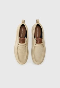 Des chaussures en suede beige avec une texture lisse, un col rembourré, et un accent en cuir marron sur la languette avec le logo Tommy Hilfiger.