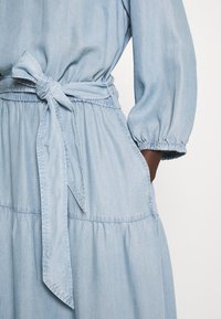 Ljusblå denimklänning med långa puffärmar, cinchad midja och en knytbälte. Har en lagerpålagd kjol och elastiska manschetter för komfort.