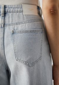 Šviesiai mėlyni džinsai, turintys galinį kišenį ir baltą prekės etiketę su įspaudu „VERO MODA“. Audinys su tekstūra ir klasikinis kirpimas.