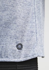 Světle modrá tkanina s texturou a mírně melírovaným vzhledem. Lem obsahuje dekorativní černou emblemu. Detail horní části oděvu.