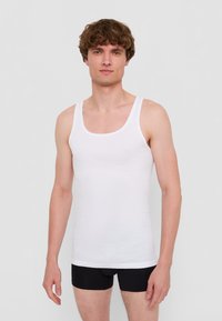 Vit bomullstopp med breda axelremmar och rund halsringning, bärs med svarta figurnära shorts. Enkelt mönster, slät textur, minimal detaljering.