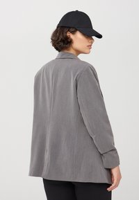 Blazer grigio con una vestibilità rilassata, design a un solo bottone e rever a punta. Cappellino nero che aggiunge un tocco informale. Tessuto morbido e forma sartoriale.