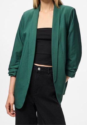 Femme portant un blazer vert foncé par-dessus un top noir tube associé à un jean noir taille haute, debout avec une main dans la poche.