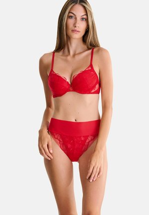 Femme aux longs cheveux portant un soutien-gorge en dentelle rouge et une culotte assortie en dentelle avec une large ceinture, debout devant un fond clair uni.