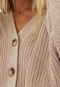 Cardigan de punto grueso beige con botones grandes, llevado por una persona con cabello rubio claro parcialmente visible.