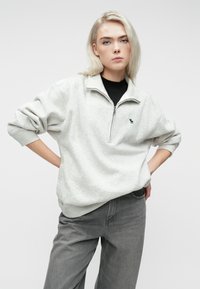 Sweat à zip gris clair en matériau doux, avec un logo vert sur la poitrine, des manches retroussées et une coupe décontractée.