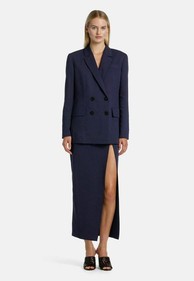 Navy double-breasted blazer with black buttons, paired with a long skirt featuring a side slit. Made from textured fabric.