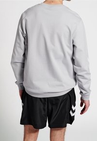 Uomo che indossa una camicia a maniche lunghe grigia chiara e pantaloni corti sportivi neri con motivi a chevron bianchi, visto da dietro su uno sfondo semplice.